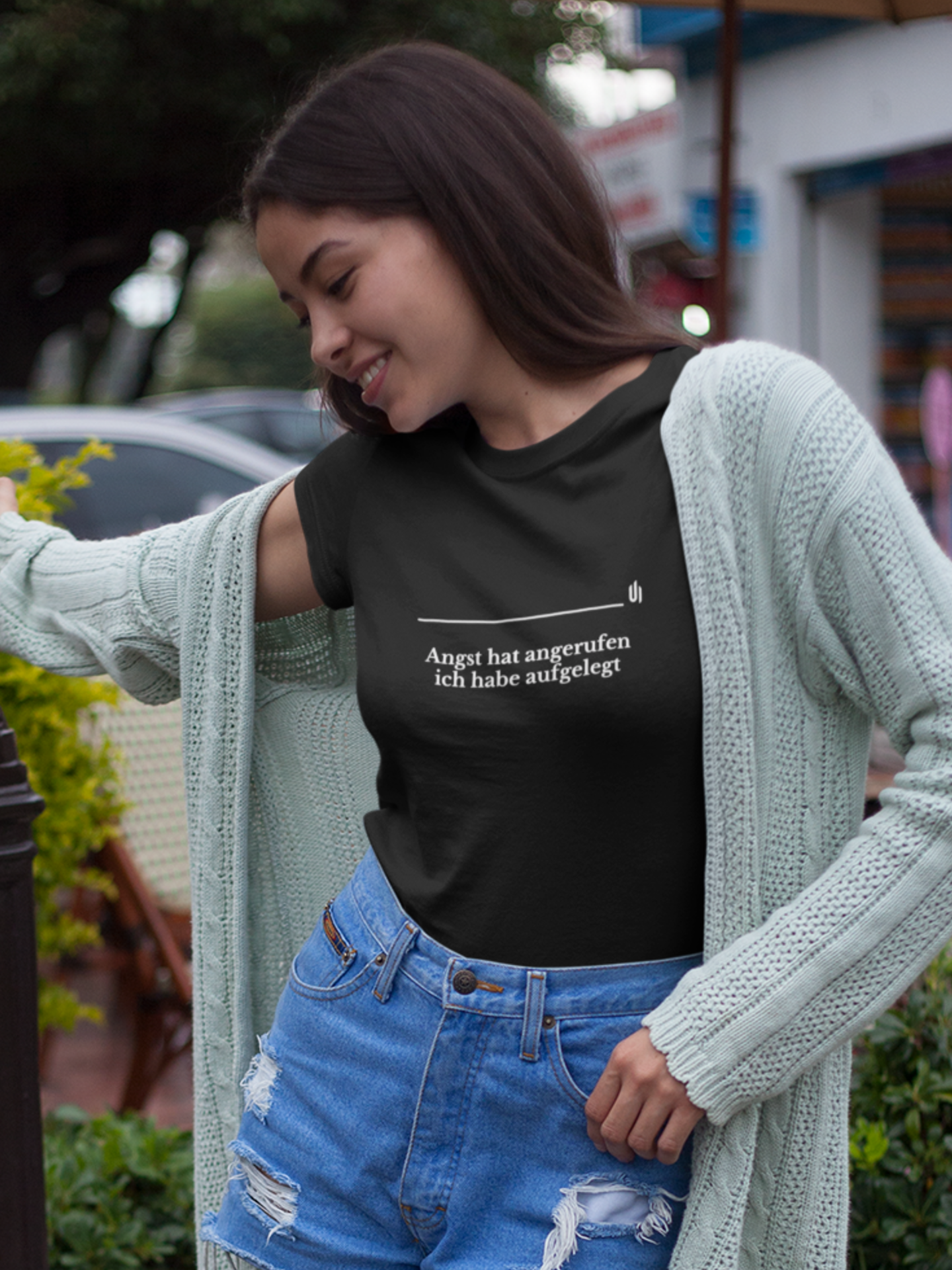 Woman wearing a black t-shirt with text, light green cardigan, and blue jeans outdoors.
