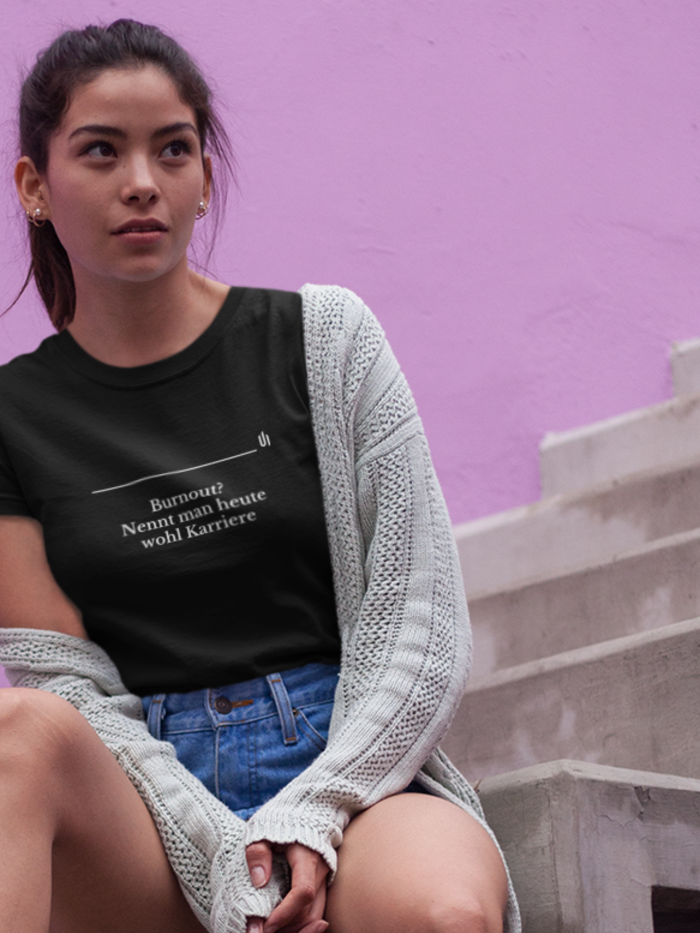 Woman wearing a black t-shirt with text Burnout_ Nennt man heute wohl Karriere - Unisex T-shirt, sitting against a pink wall.
